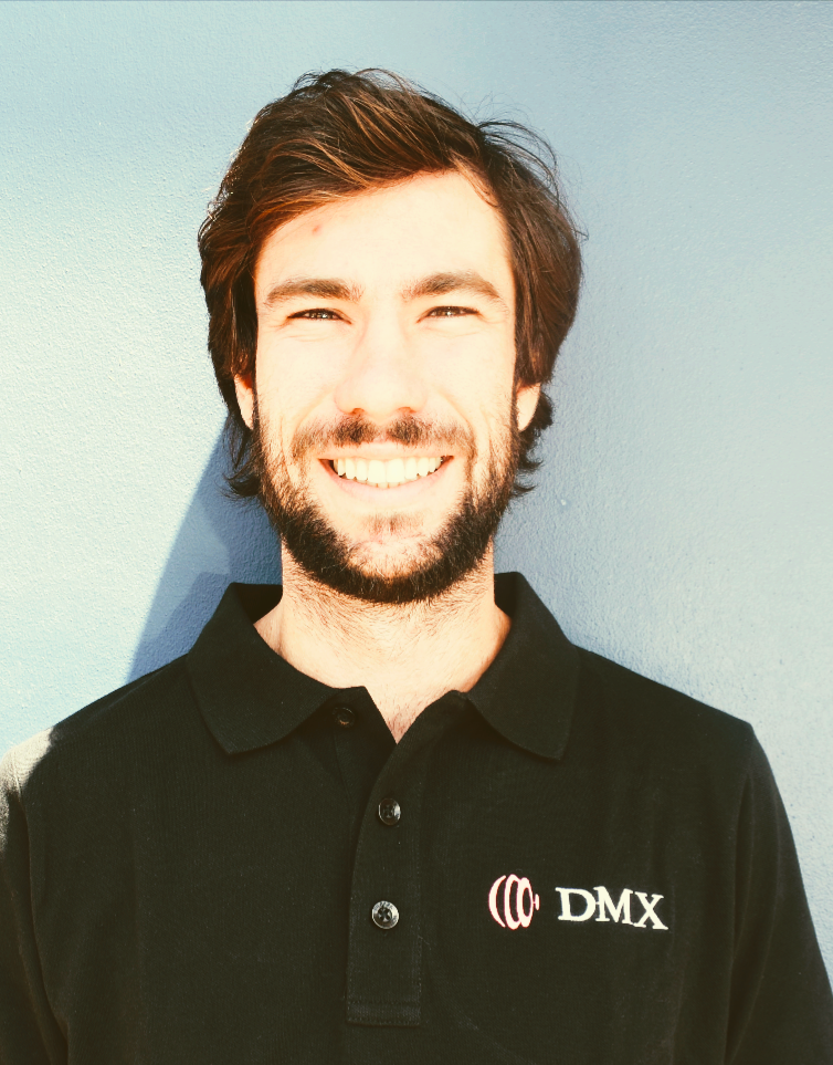 Smiling man in a black DMX polo shirt against a light blue wall.