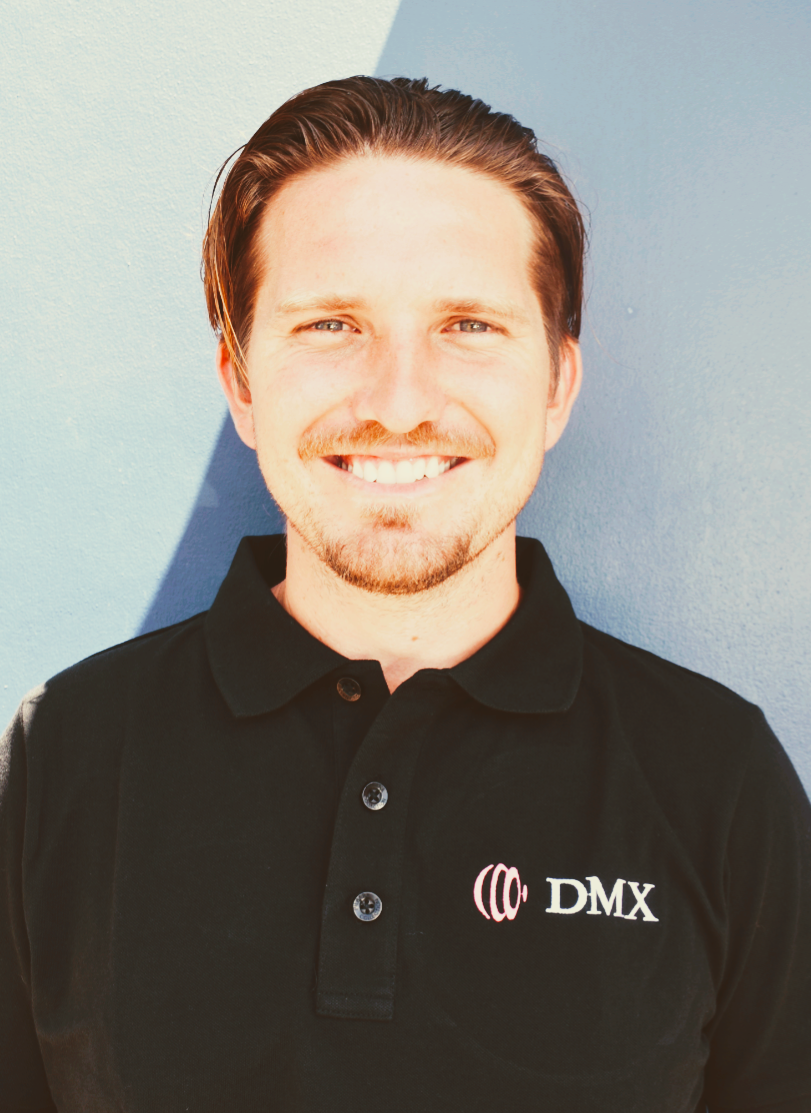 Smiling person in a black polo shirt with a DMX logo, standing against a blue wall.