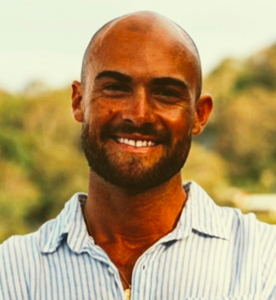 Smiling bald man with a beard wearing a striped shirt outdoors, with a blurred natural background