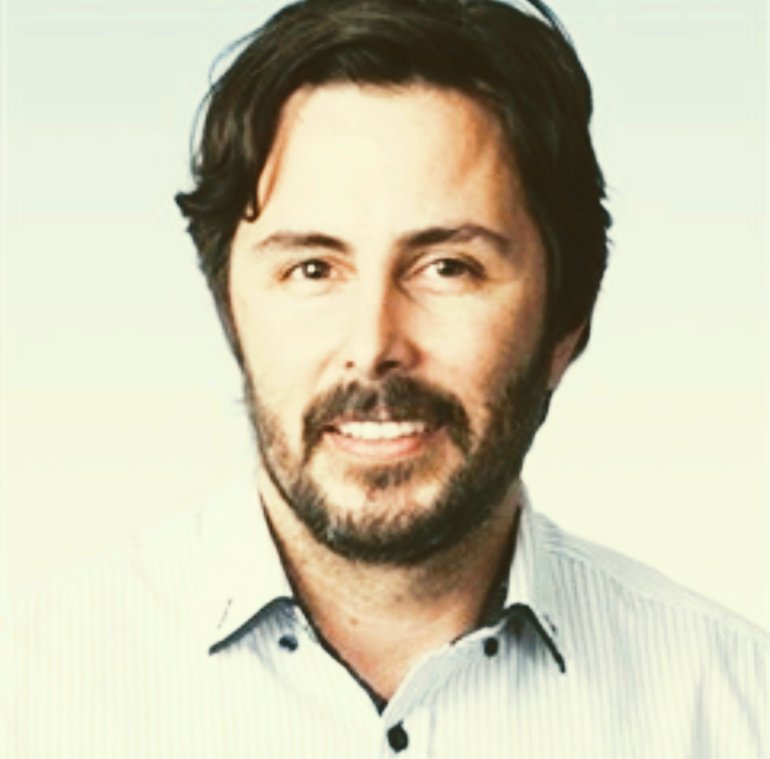 Portrait of a smiling man with dark hair and beard, wearing a light blue collared shirt on a pale background