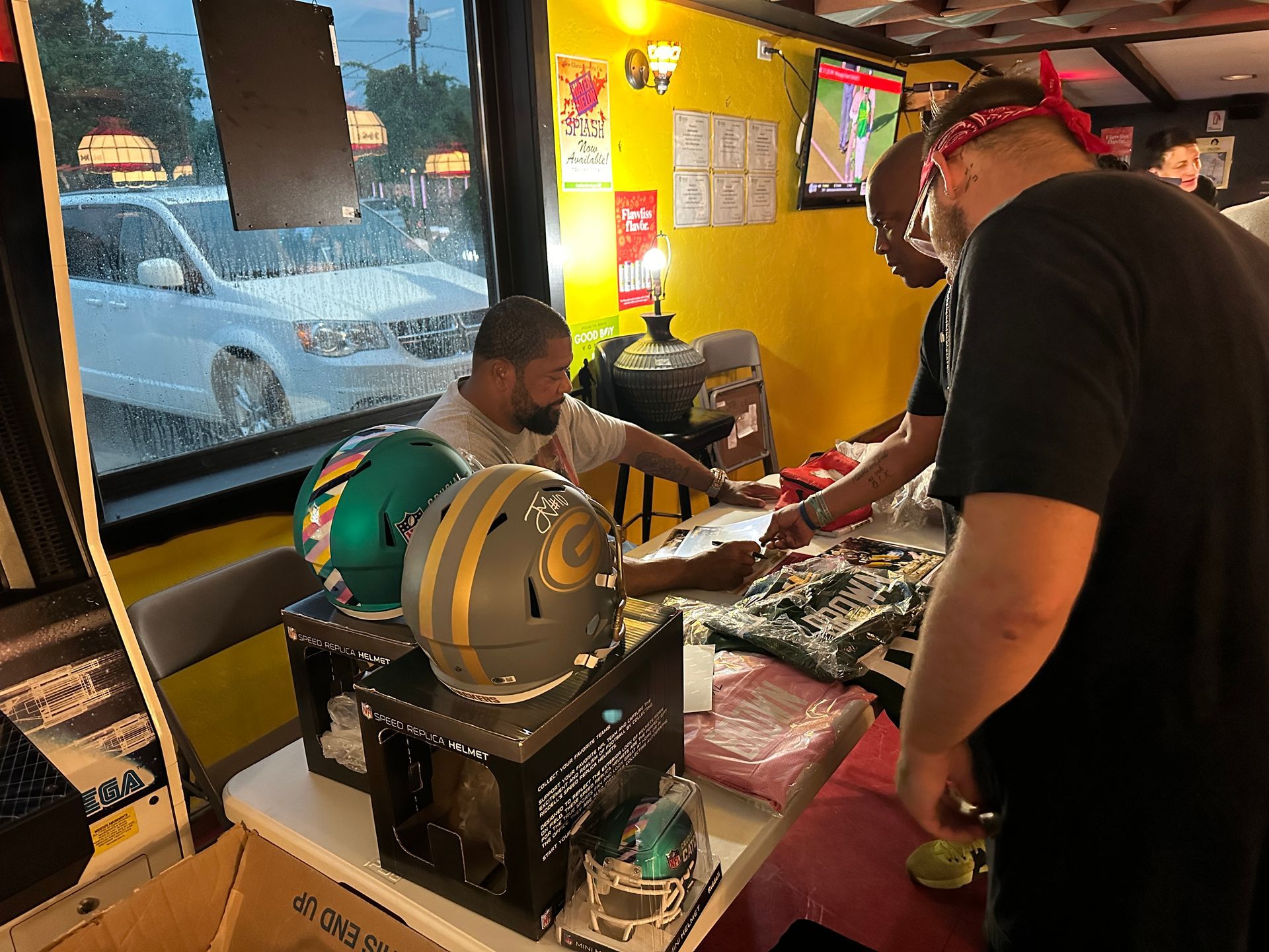 People examining sports merchandise inside a store, including football helmets, with a car visible outside.