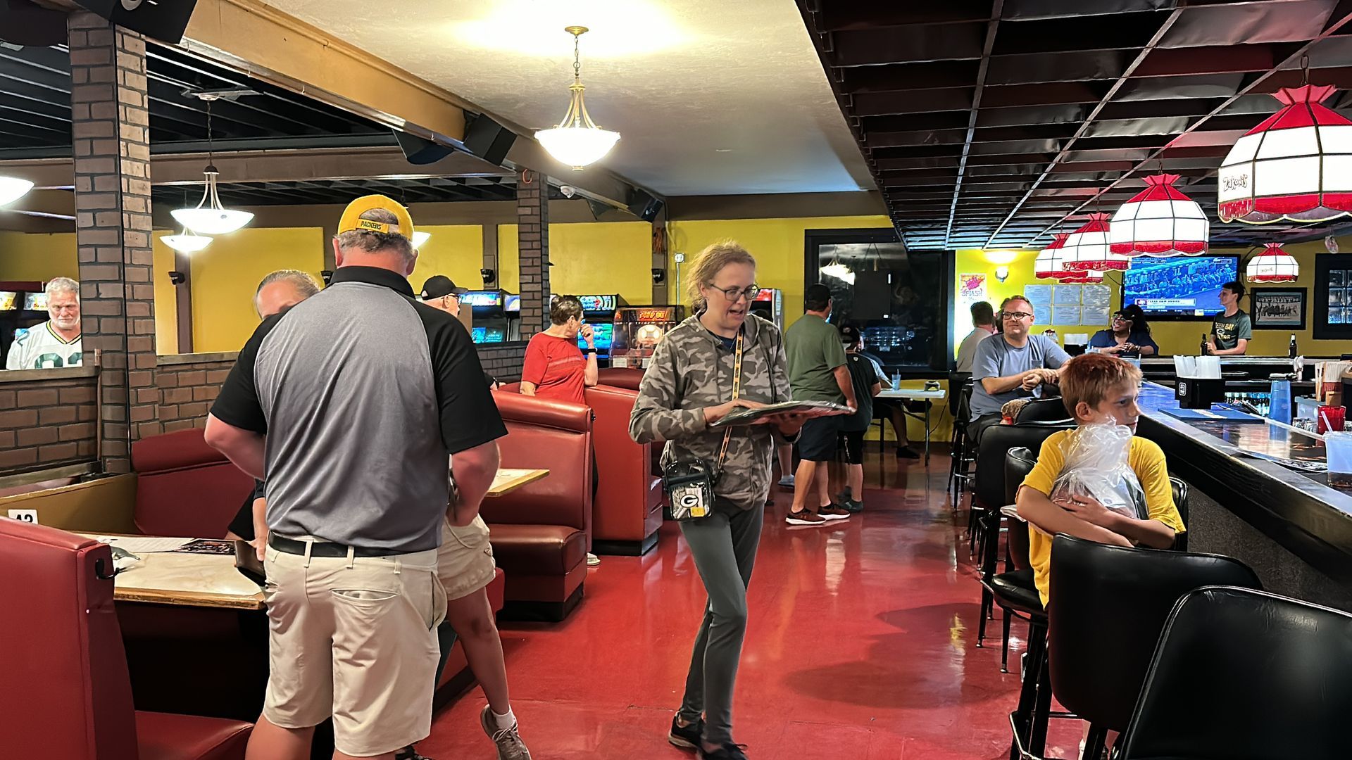 People in a diner. A server holds a tray. Red floor, booths, bar, TVs, and arcade games.