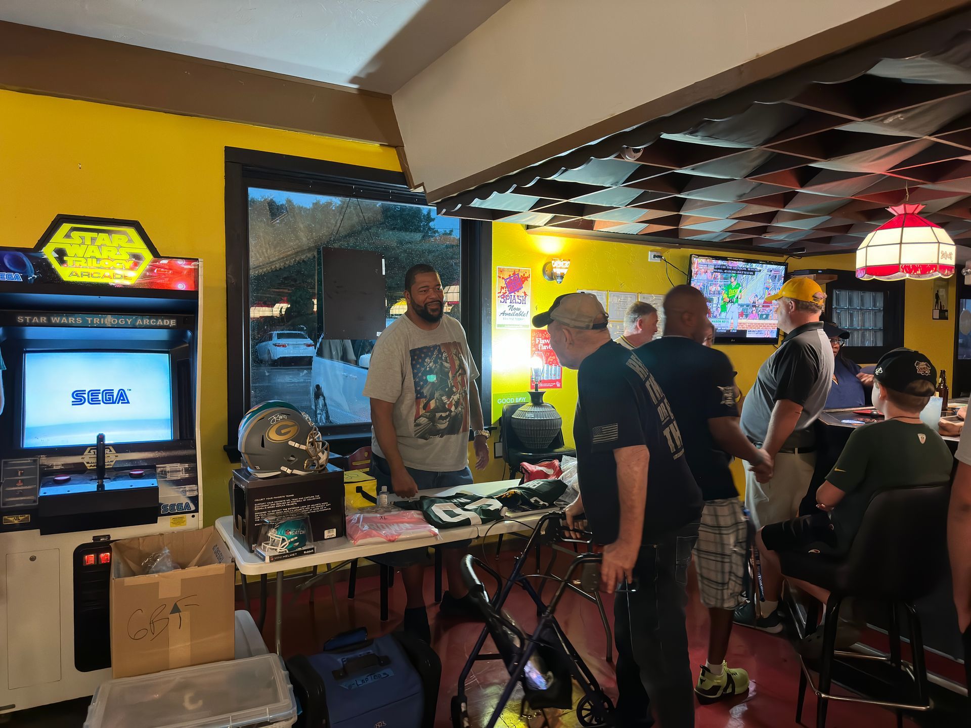 People at a retro game event; arcade cabinet, tables, people, indoor setting.