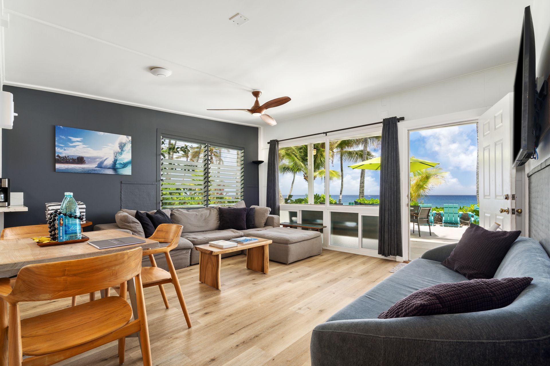 Living room with ocean view, gray sectional, wooden table & chairs, open door to patio.