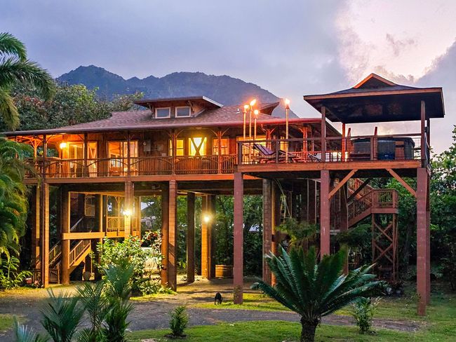 Wooden stilt house with warm lighting, surrounded by lush greenery, under a twilight sky.