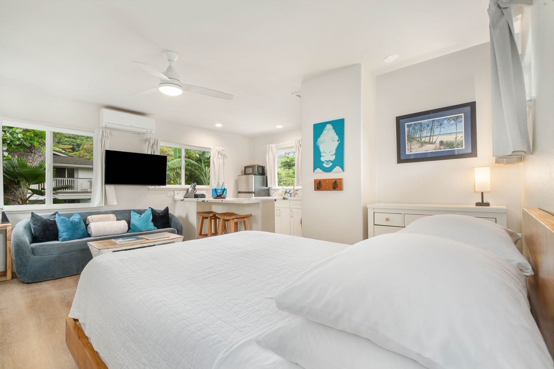 Bed in foreground, living area with sofa & TV, kitchen in background, white walls, wood floors.