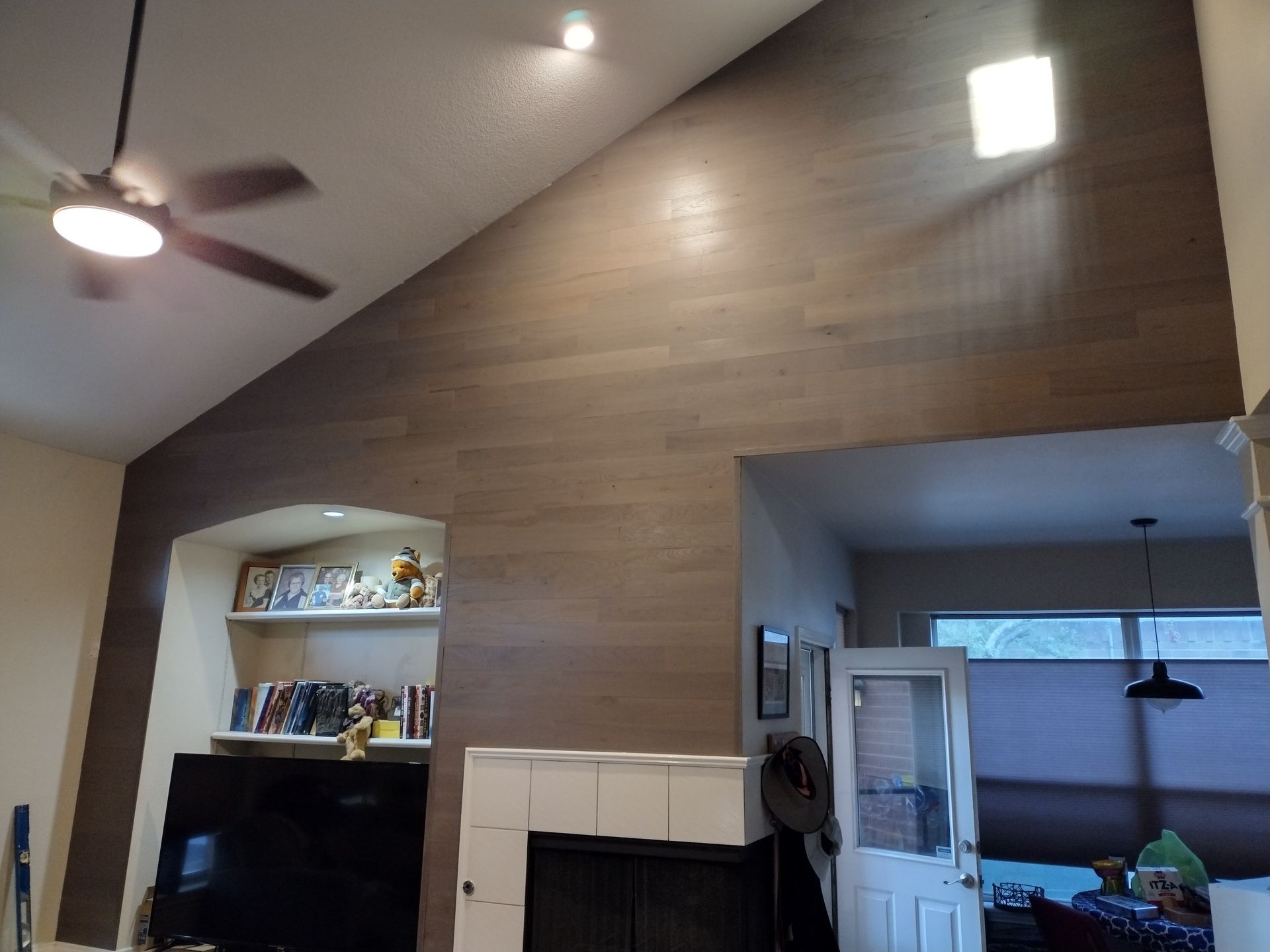 Living room with high wood-paneled wall, built-in shelves, fireplace, ceiling fan, and window with blinds.