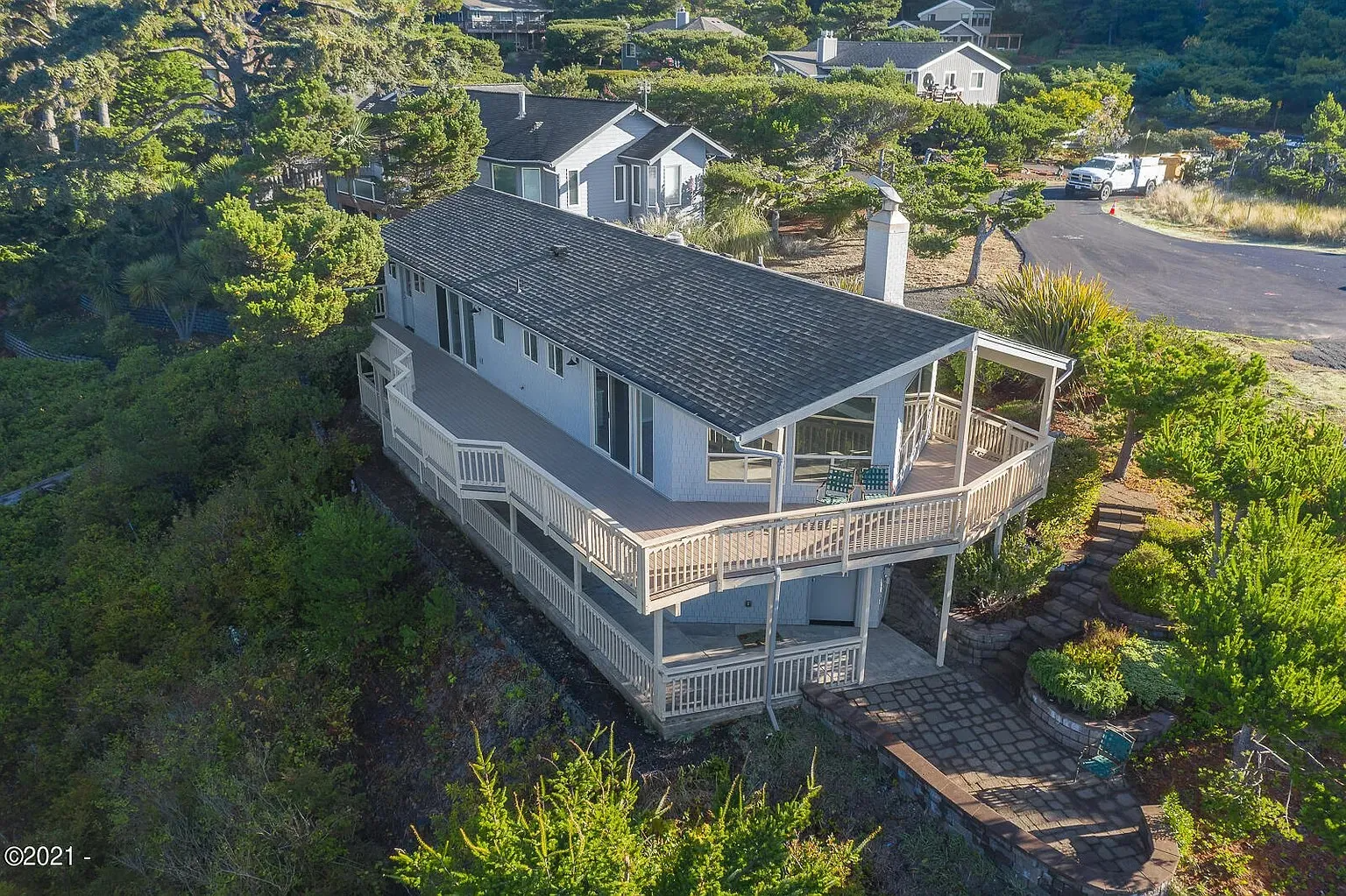 Aerial view of a gray house with a wraparound deck on a wooded hillside, near neighboring homes.