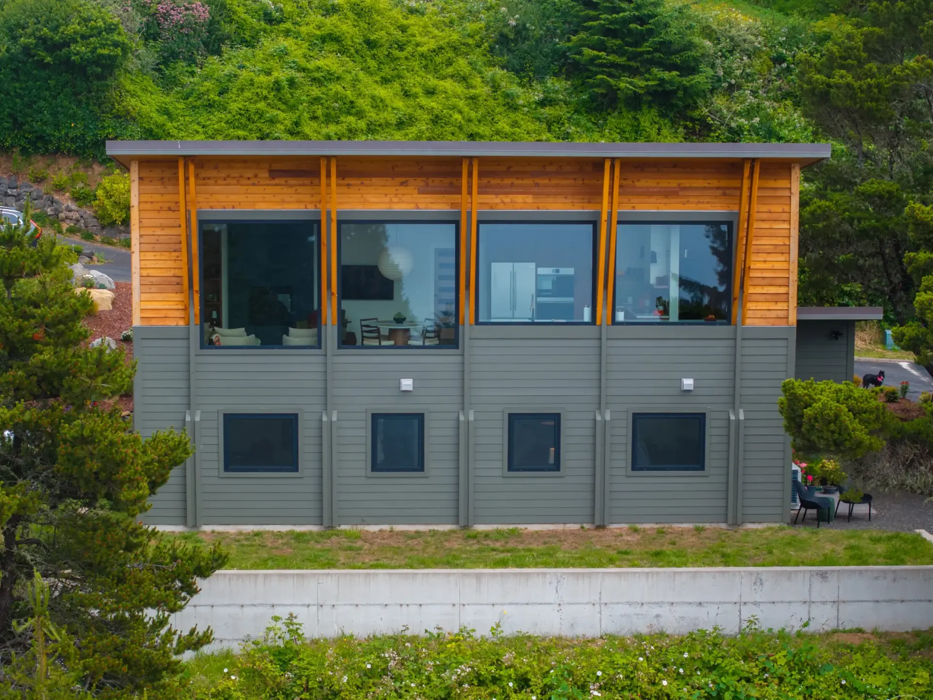 Modern two-story gray house with wood accents set against a green hillside