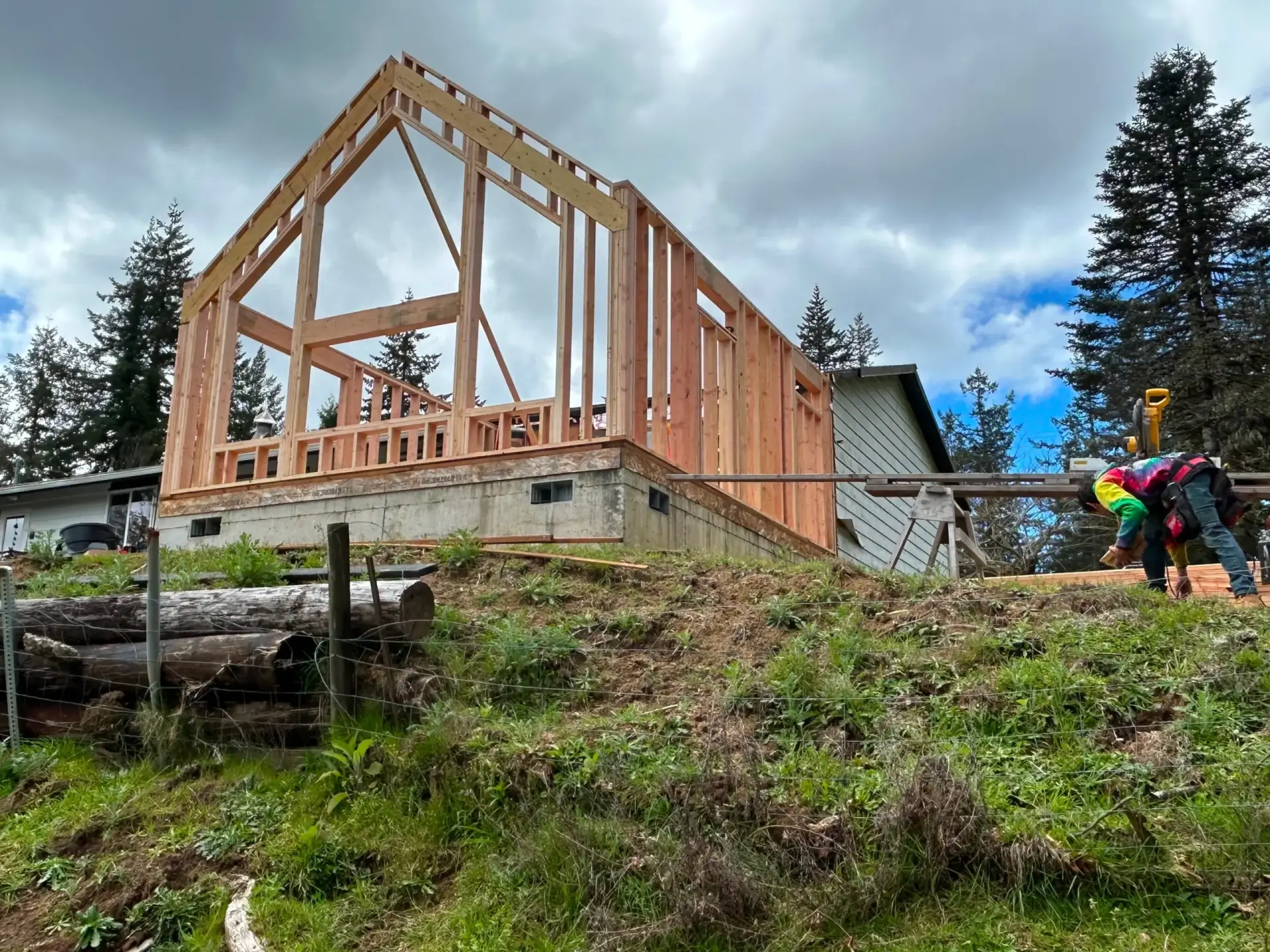 Wood-framed house under construction on a sloped lot with workers and cloudy sky