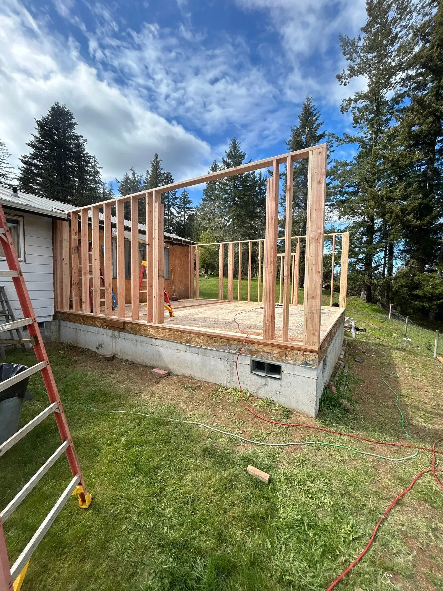 Wooden house frame under construction on a concrete foundation, with a ladder and grassy yard under a blue sky