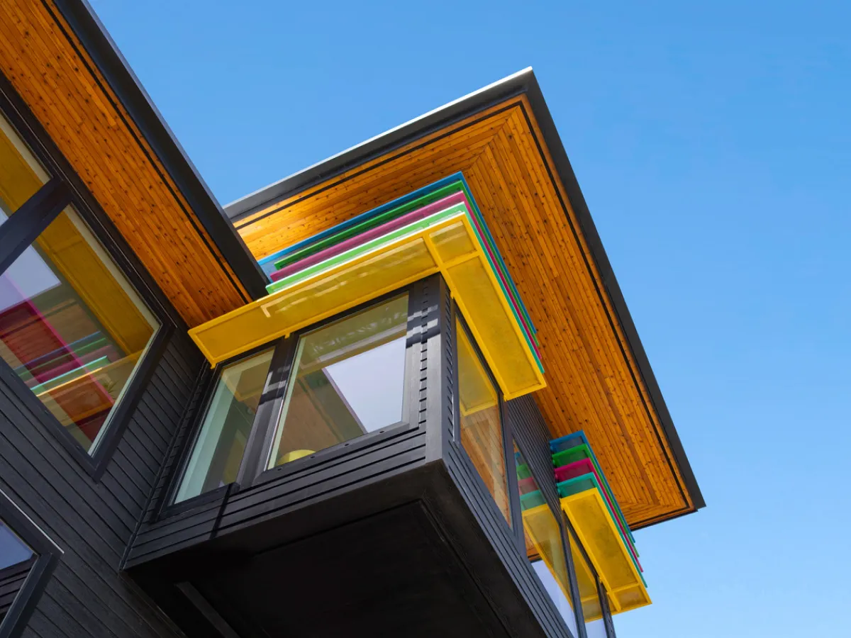 Modern house corner with black siding, yellow trim, and colorful striped roof accents against a blue sky