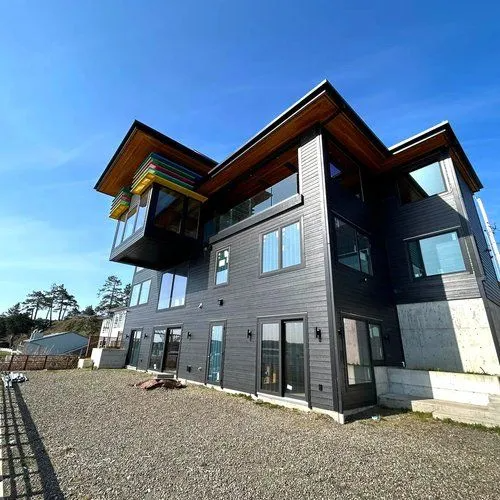 Modern dark-gray house with wood-trimmed roof and large windows on a gravel lot under a blue sky