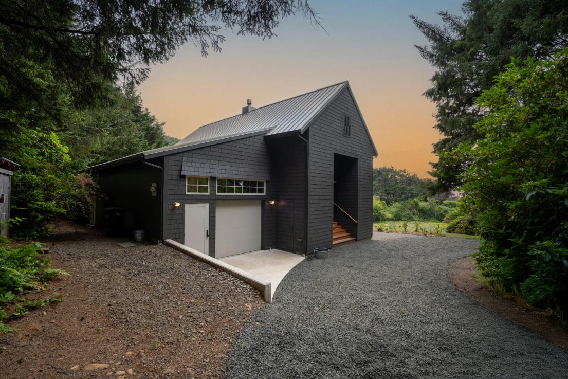 Modern dark-gray cabin with a gravel driveway, surrounded by trees at dusk