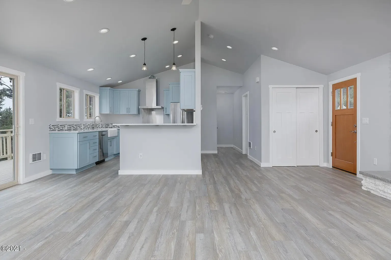 Bright open kitchen and living area with gray floors, white walls, recessed lights, and a wood front door