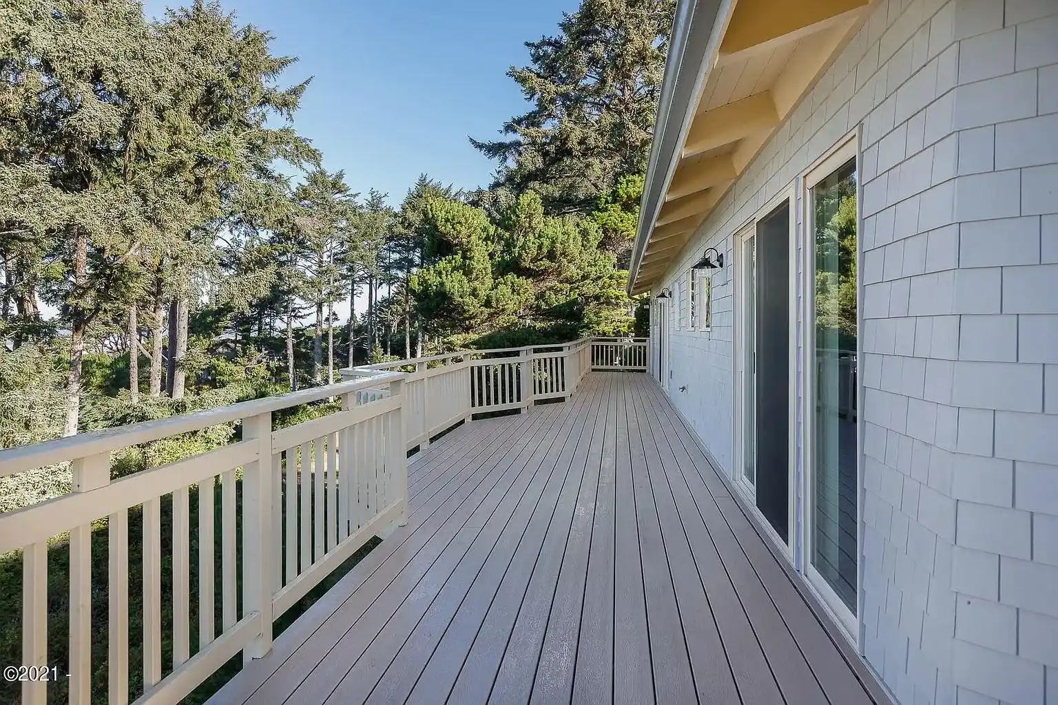 Wide wooden deck with white railing beside a house, overlooking trees and greenery