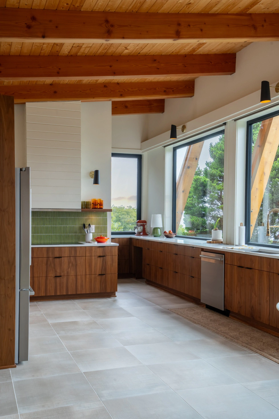 Modern kitchen with wood cabinets, white tile floor, exposed beams, and large windows overlooking greenery