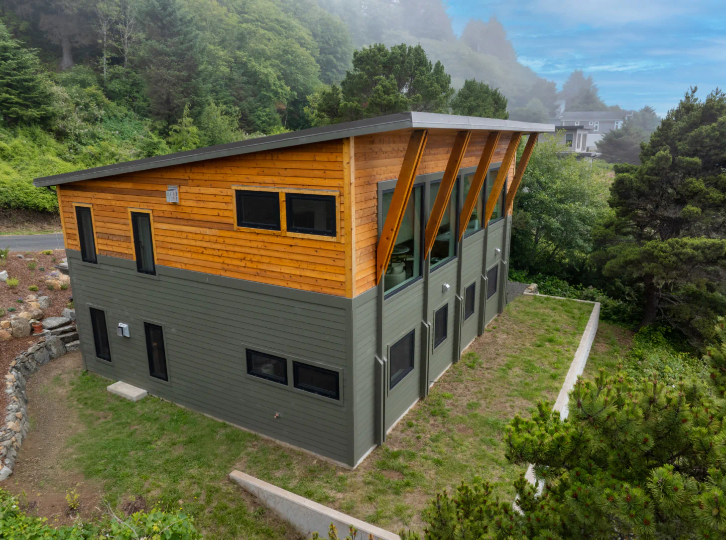 Modern two-story wooden and gray building on a hillside, surrounded by lush greenery and misty mountains