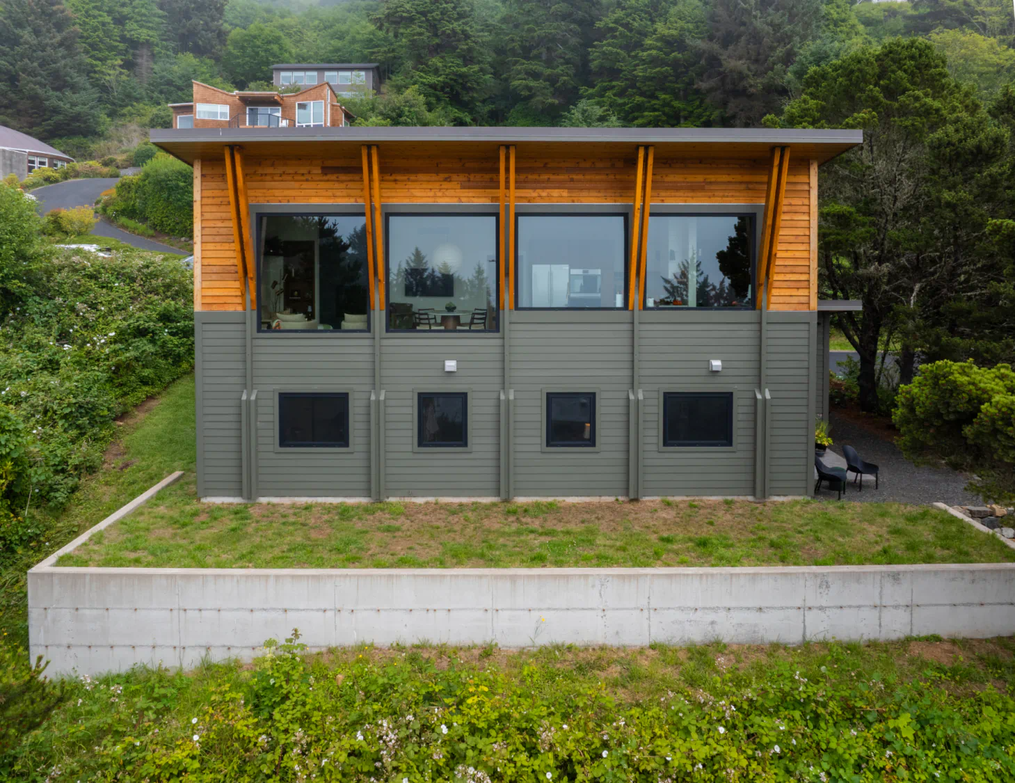Modern two-story house with wood and gray siding, large windows, and a flat roof on a grassy hillside