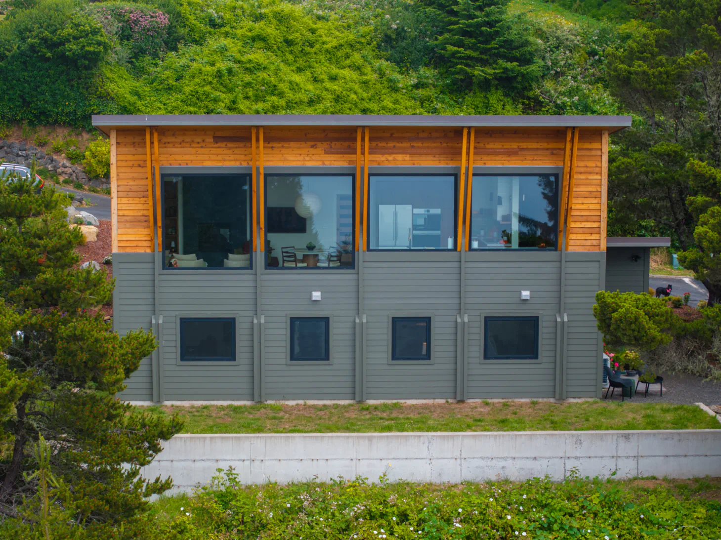 Modern gray-and-wood house on a hillside with large windows and lush greenery behind it