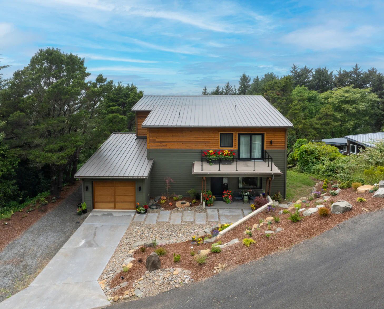 Modern two-story house with garage, gray roof, and stone landscaping on a sloped wooded lot