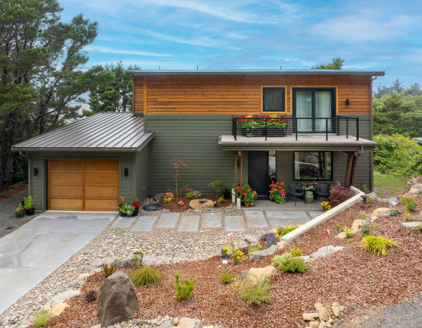 Modern two-story house with garage, wood siding, balcony, and landscaped front yard under a blue sky