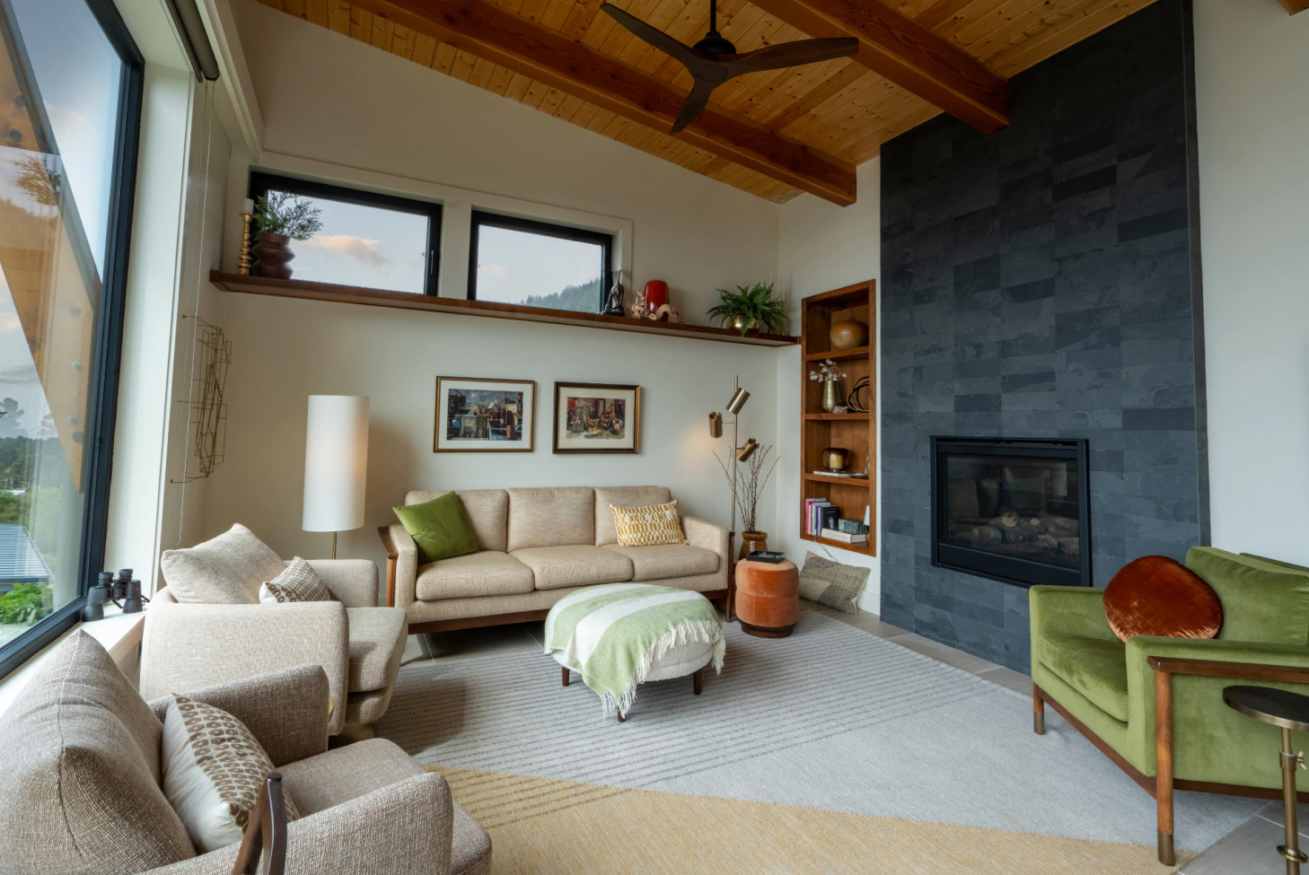 Cozy living room with beige sofa, green chair, large windows, and dark stone fireplace with wood ceiling