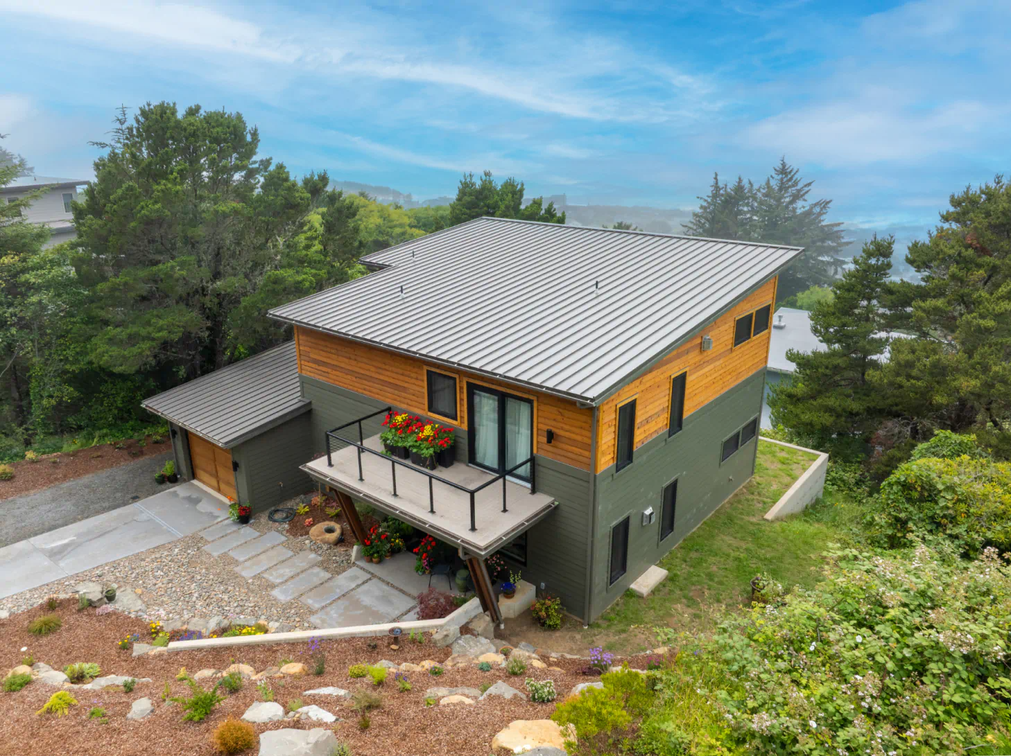 A modern two-story house with gray roof, wood siding, and a balcony surrounded by trees and greenery.
