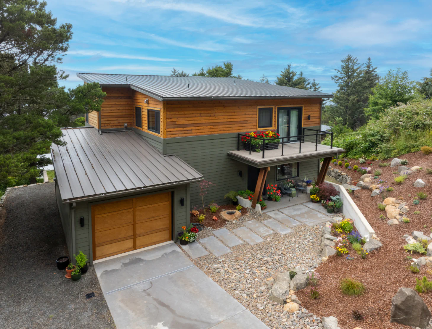 Modern wood-and-stone house with metal roof, balcony, and landscaped front yard on a hillside.