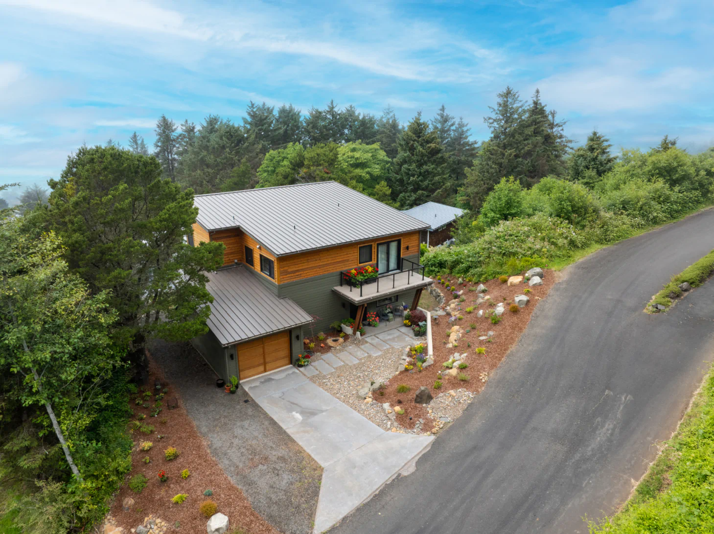 Aerial view of a modern house with a silver roof beside a curved road, surrounded by trees and greenery