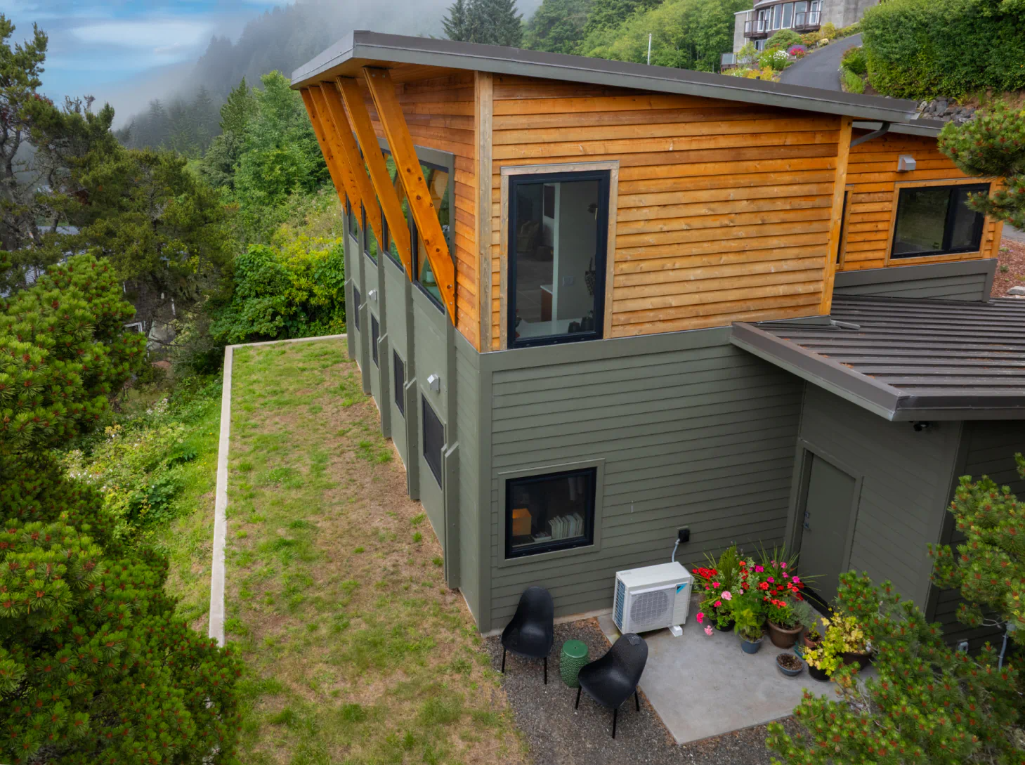 Modern hillside house with wood siding, gray lower walls, and a small patio overlooking greenery