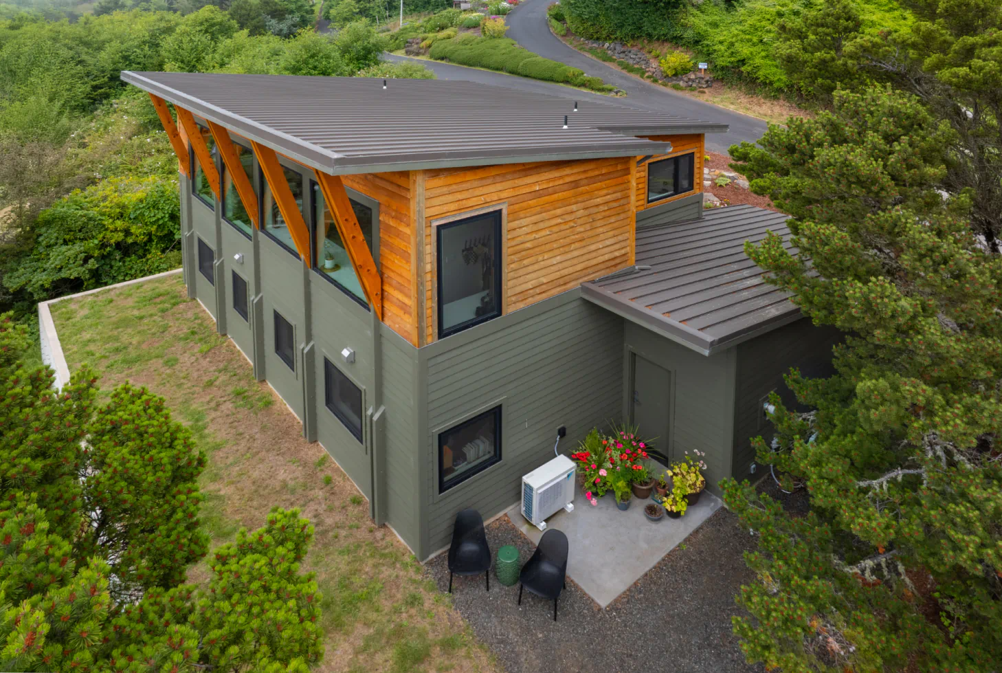 Aerial view of a modern gray house with wood siding, solar panels, and a small patio surrounded by greenery