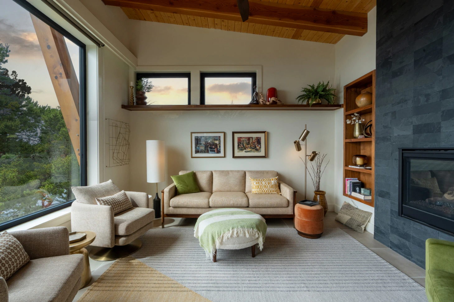 Modern living room with beige sofa, armchairs, green ottoman, large windows, and a dark stone fireplace.