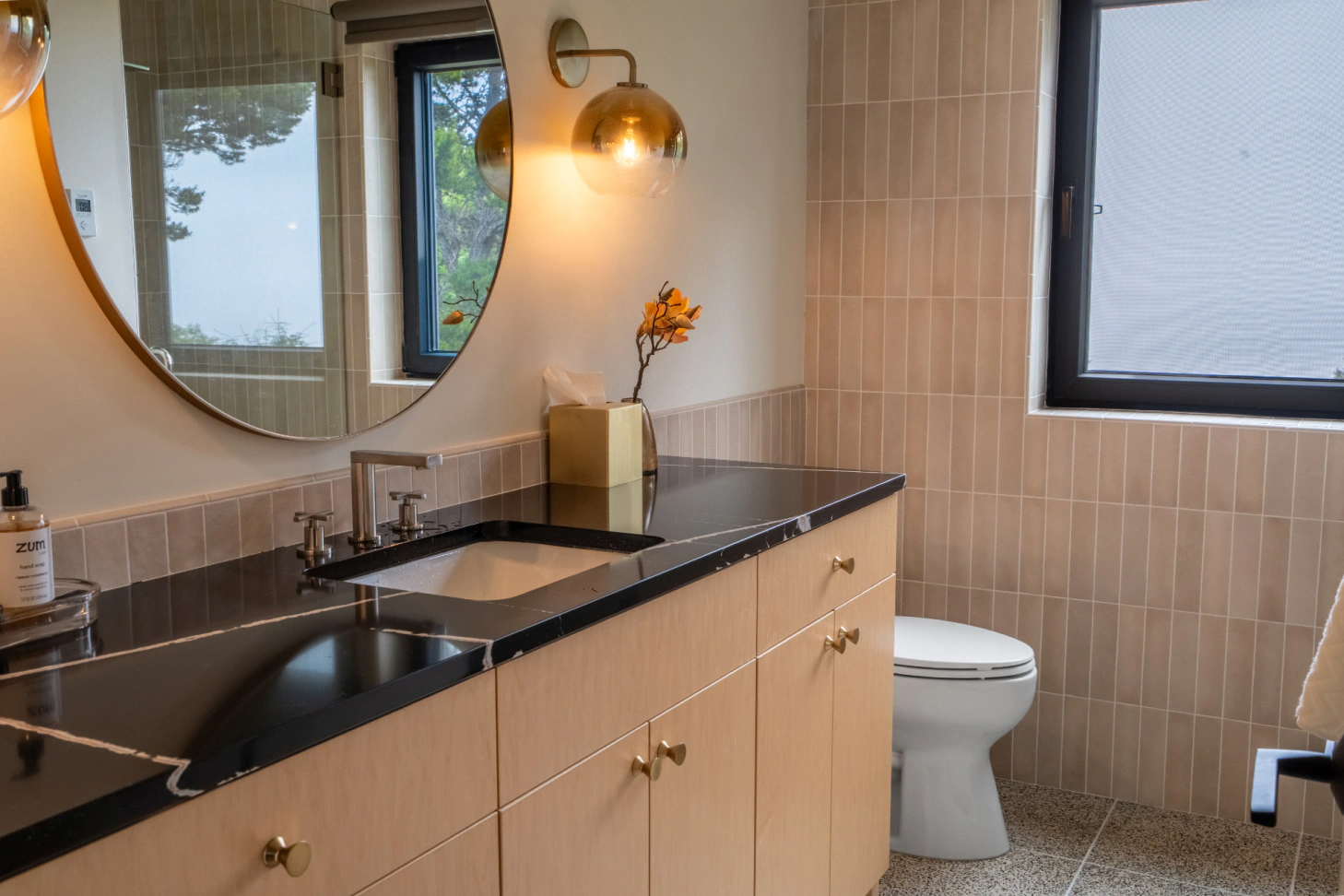 Modern bathroom with vanity, black countertop, round mirror, wall sconce, toilet, and tiled walls.