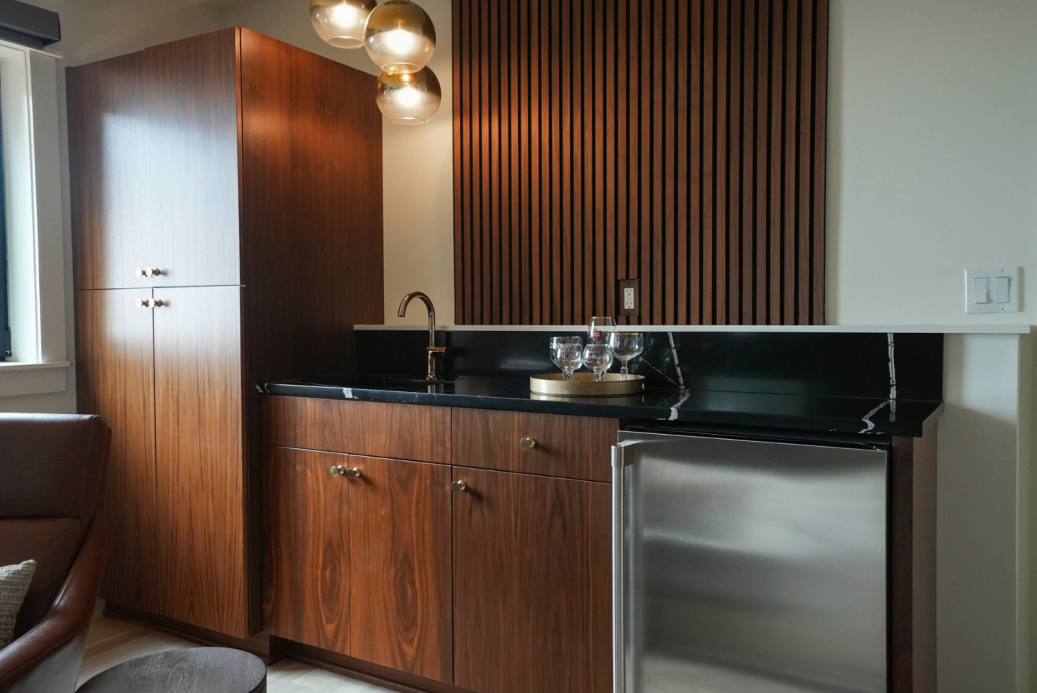 Modern kitchenette with dark wood cabinets, black countertop, stainless mini-fridge, and warm pendant lights