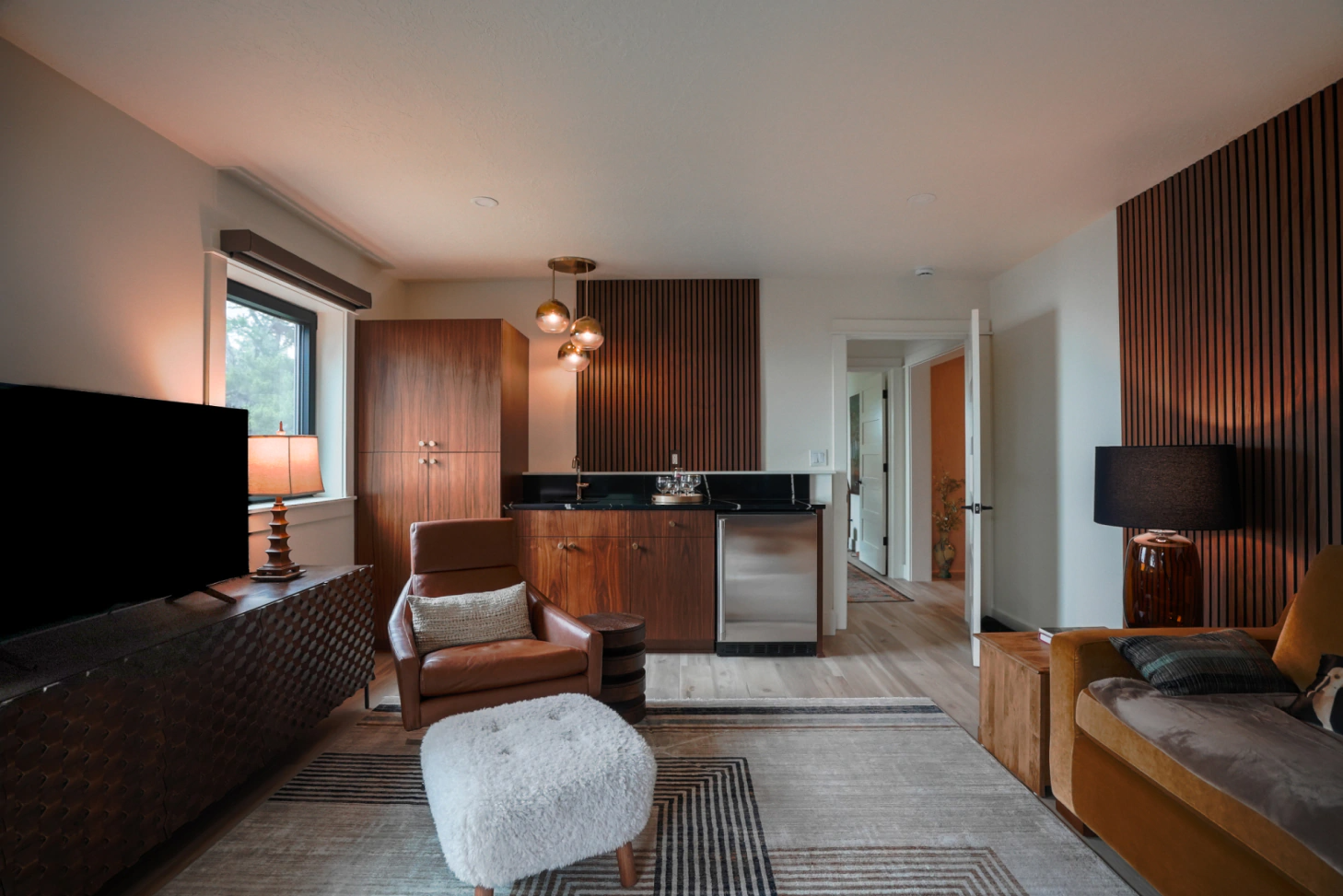 Modern living room with wood accents, sofa, ottoman, TV, and small kitchen area lit by warm lamps