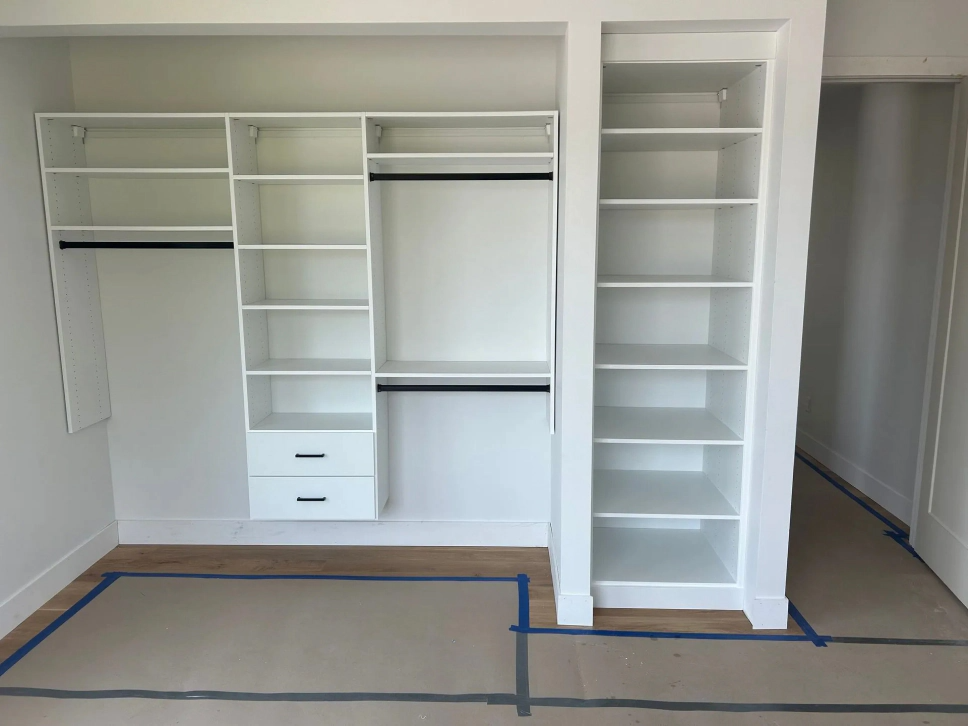 Empty bedroom closet with white built-in shelving, hanging rods, and drawers, marked on the floor with blue tape.