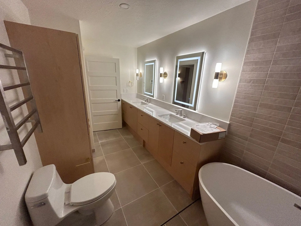 Modern bathroom with double vanity, illuminated mirrors, toilet, tub, and beige tile floors