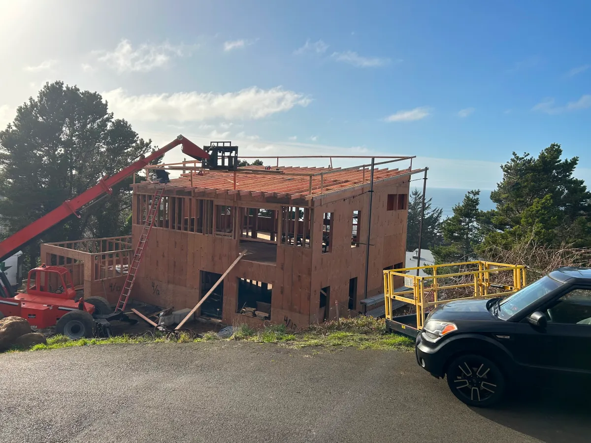 Wood-framed house under construction with a crane, on a hillside near parked vehicles and trees