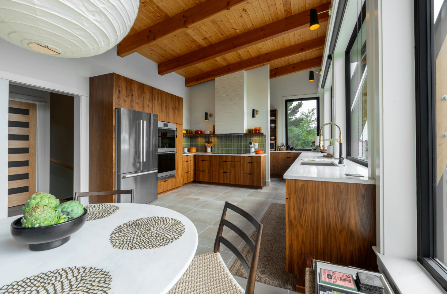 Modern kitchen with stainless steel appliances, wood cabinetry, exposed beam ceiling, and a dining table by large windows
