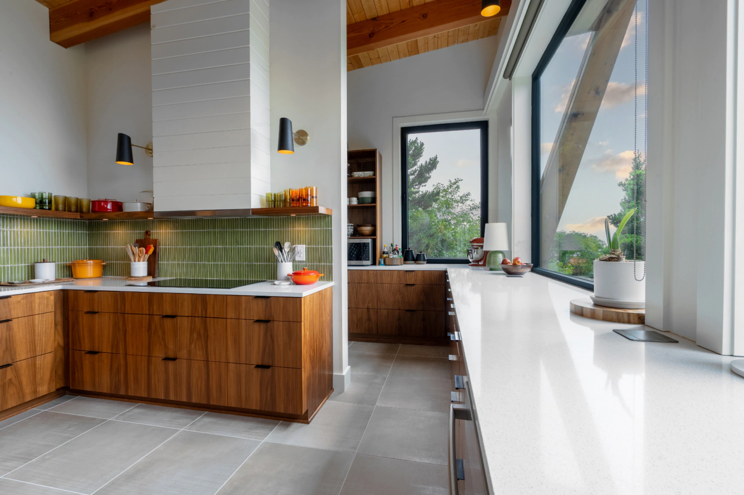 Modern kitchen with wood cabinets, green tile backsplash, and a long white countertop by large windows.