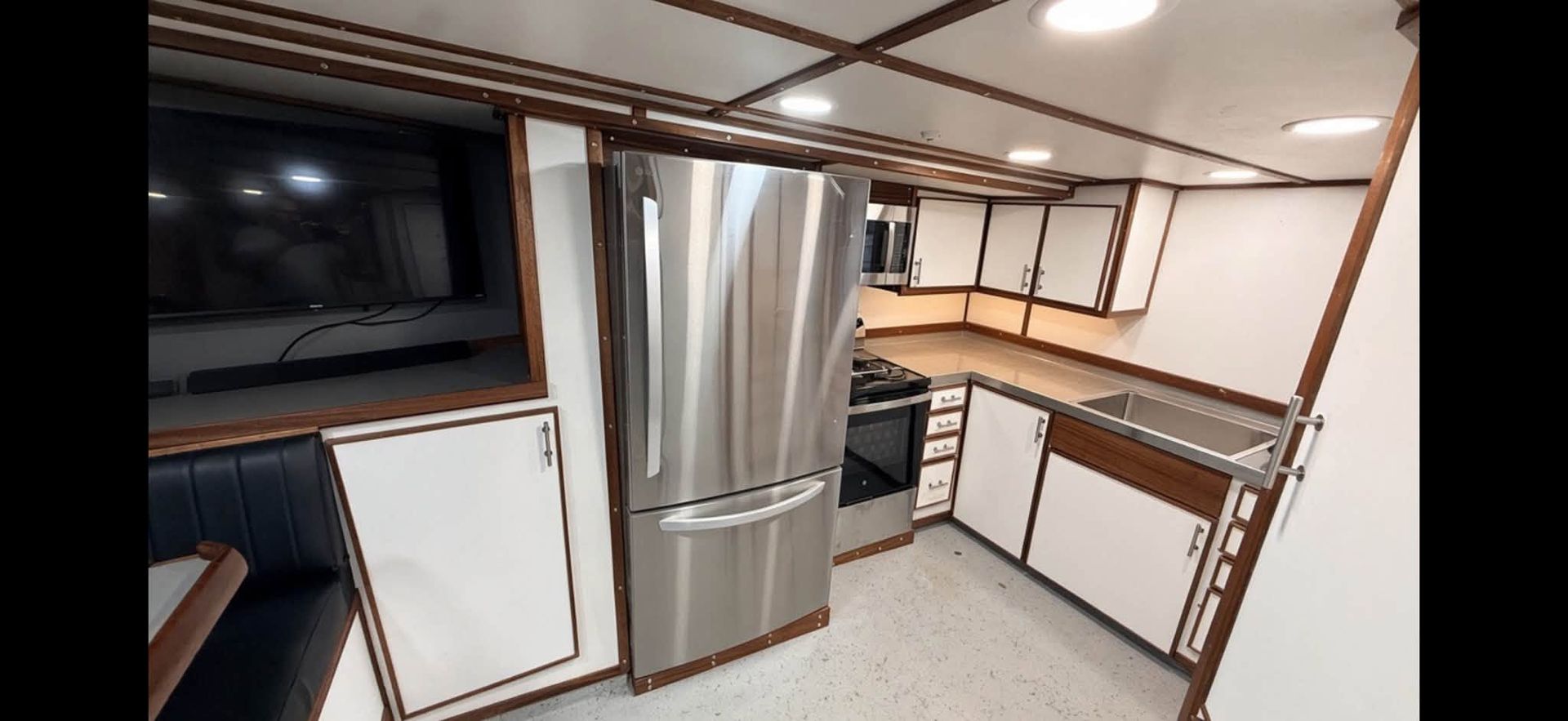 Compact kitchen with stainless steel fridge, oven, sink, white cabinets, and built-in TV