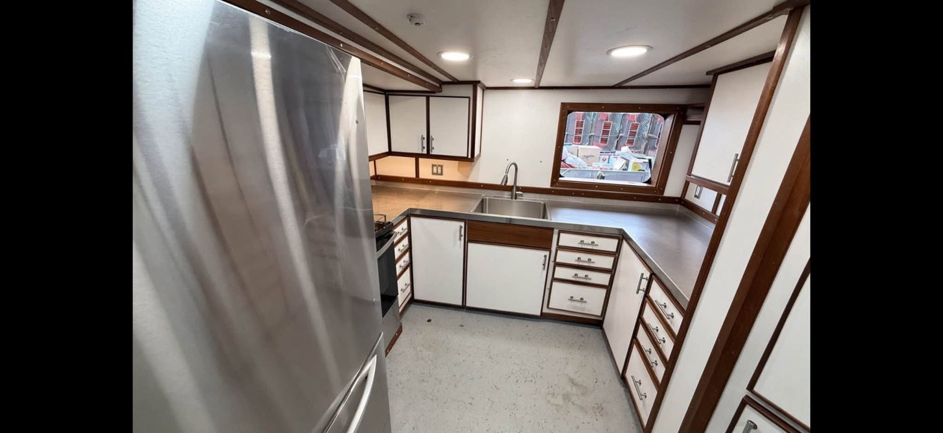 Compact kitchen interior with stainless fridge, white cabinets, wood trim, and windowed counter area