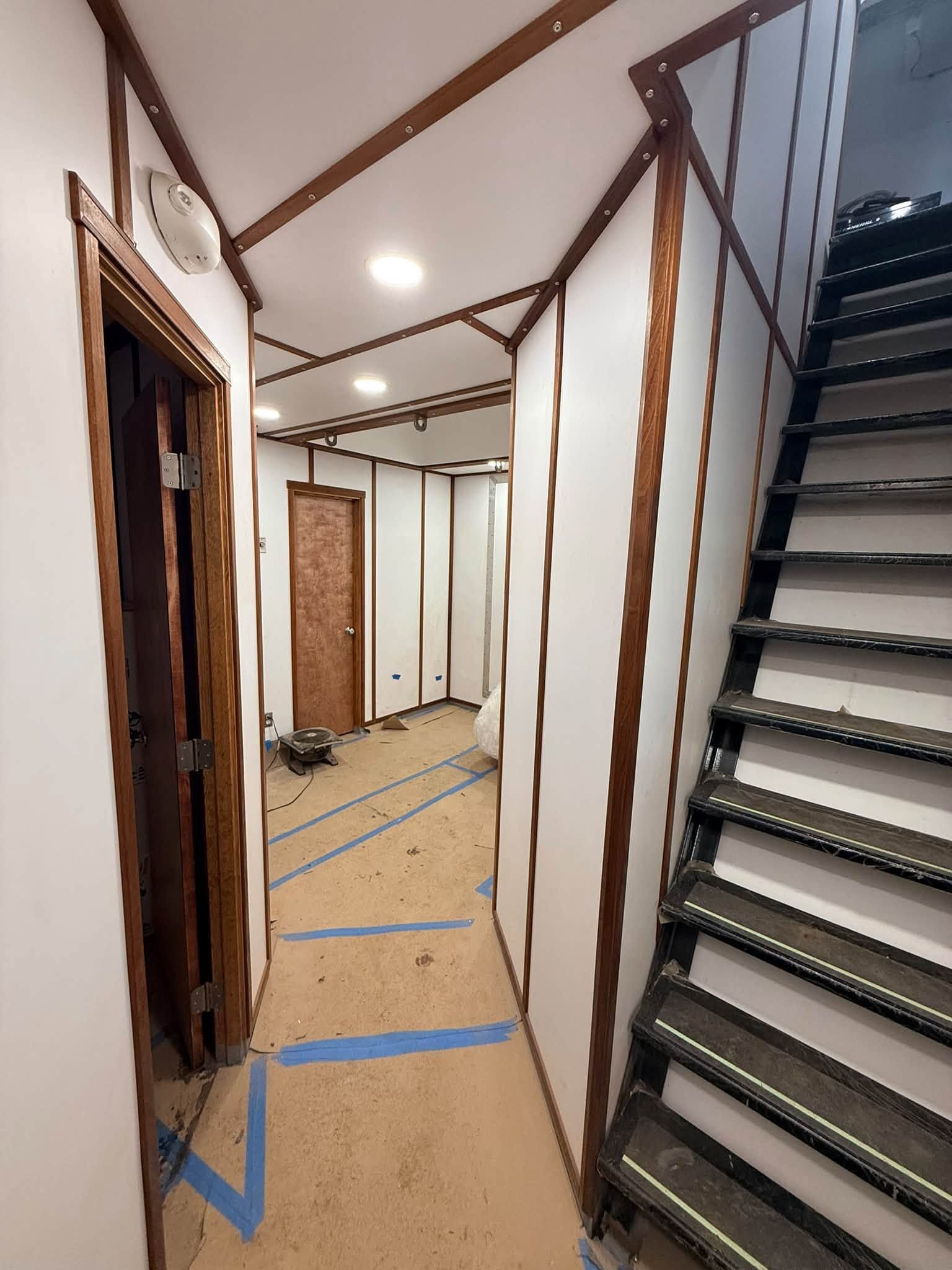 Narrow hallway in a house under renovation, with taped floors, exposed beams, and stairs on the right.