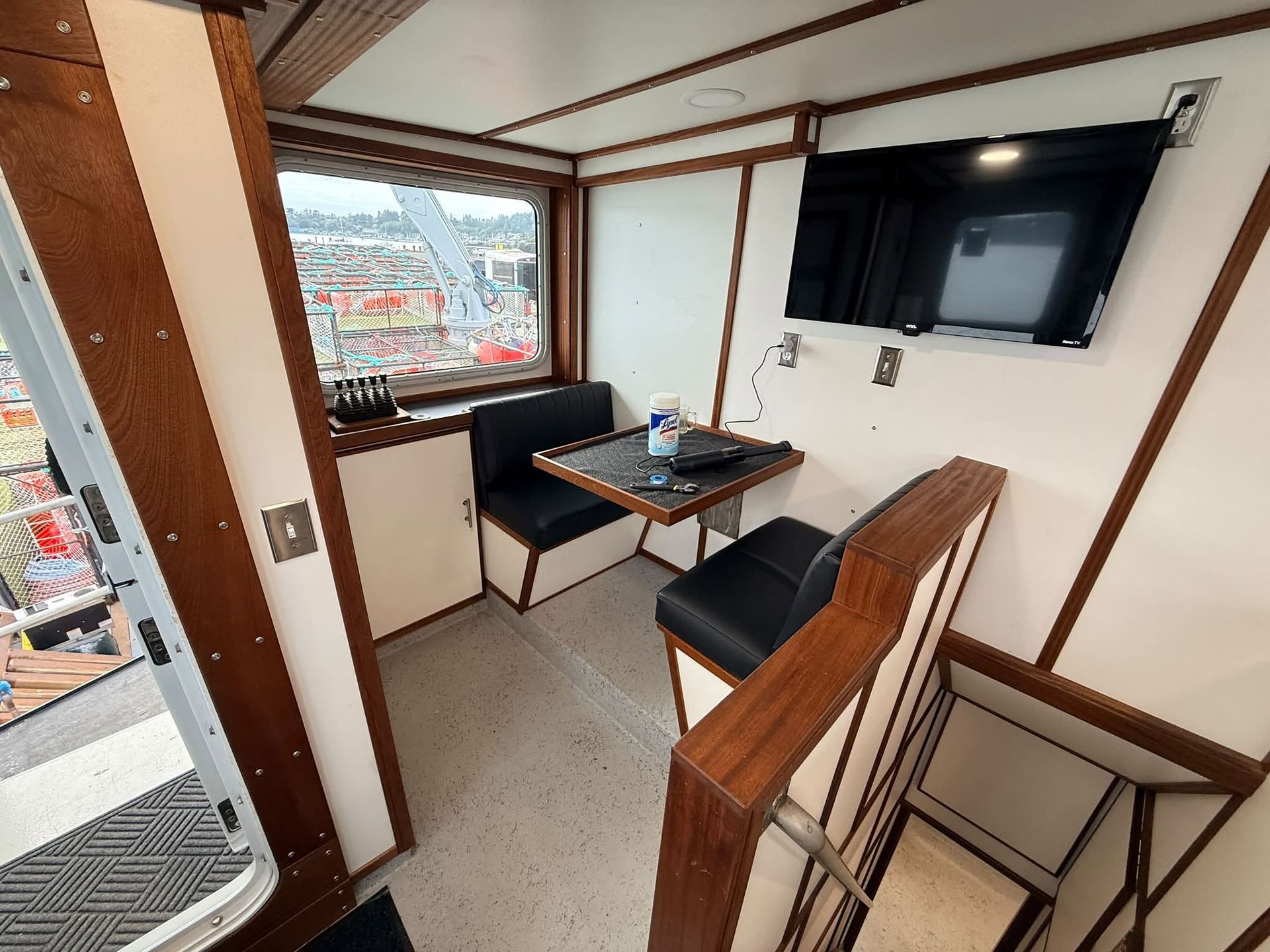 Small cabin lounge with two black chairs, a table, wall-mounted TV, and a harbor view through the window