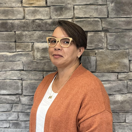 A woman wearing glasses and an orange sweater is standing in front of a brick wall.
