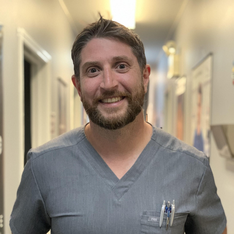 A man with a beard is wearing a scrub top and smiling.