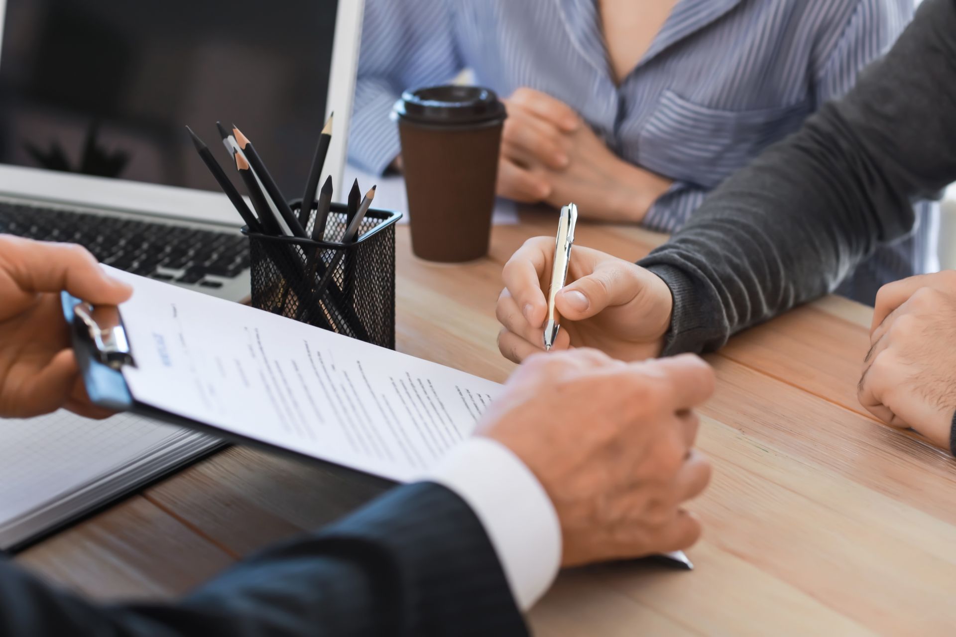 Hands signing a document at a meeting table with a coffee cup and laptop nearby