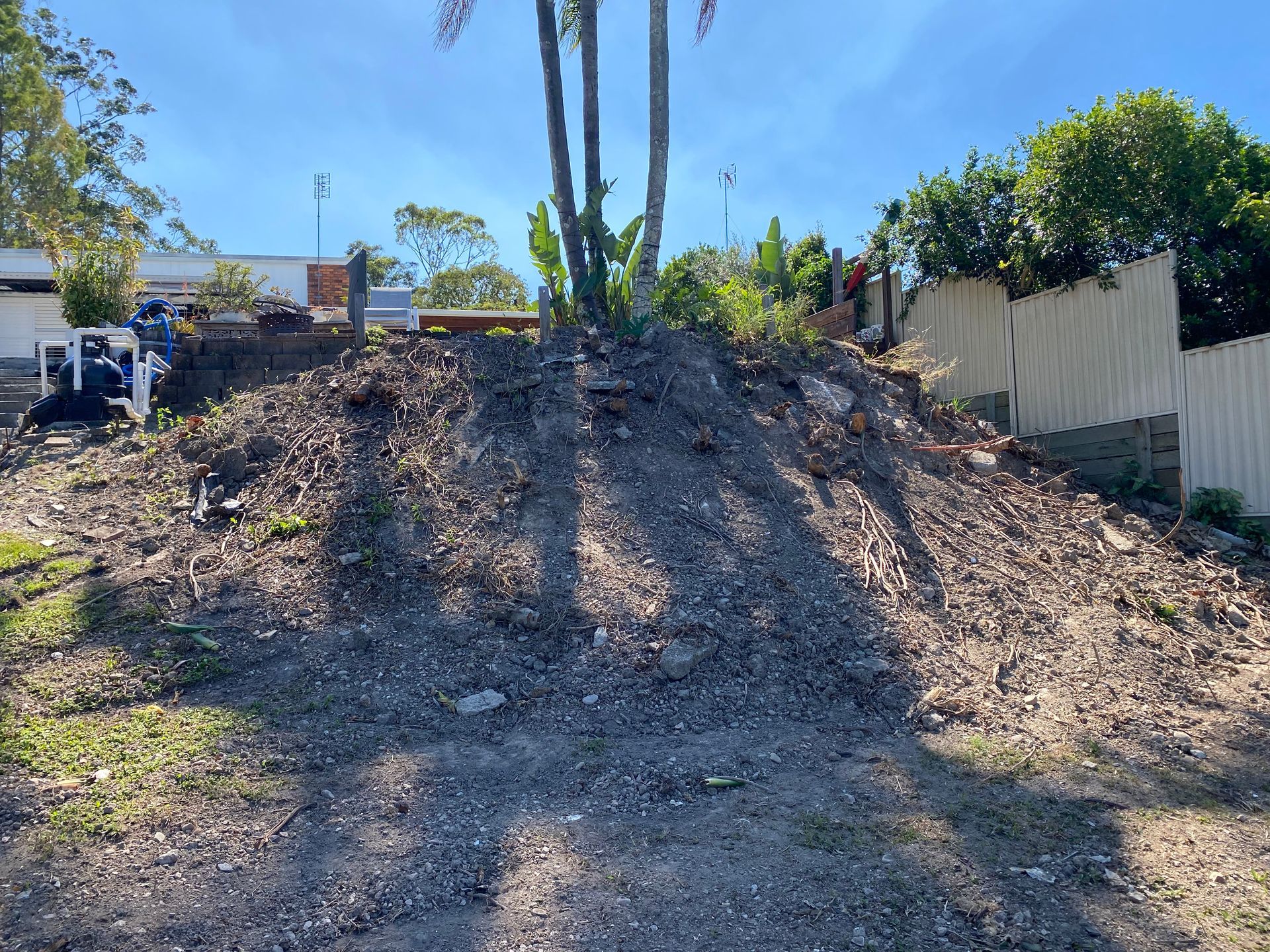 Pile of dirt, rocks, and debris; residential area. Overcast sky and green trees in background.
