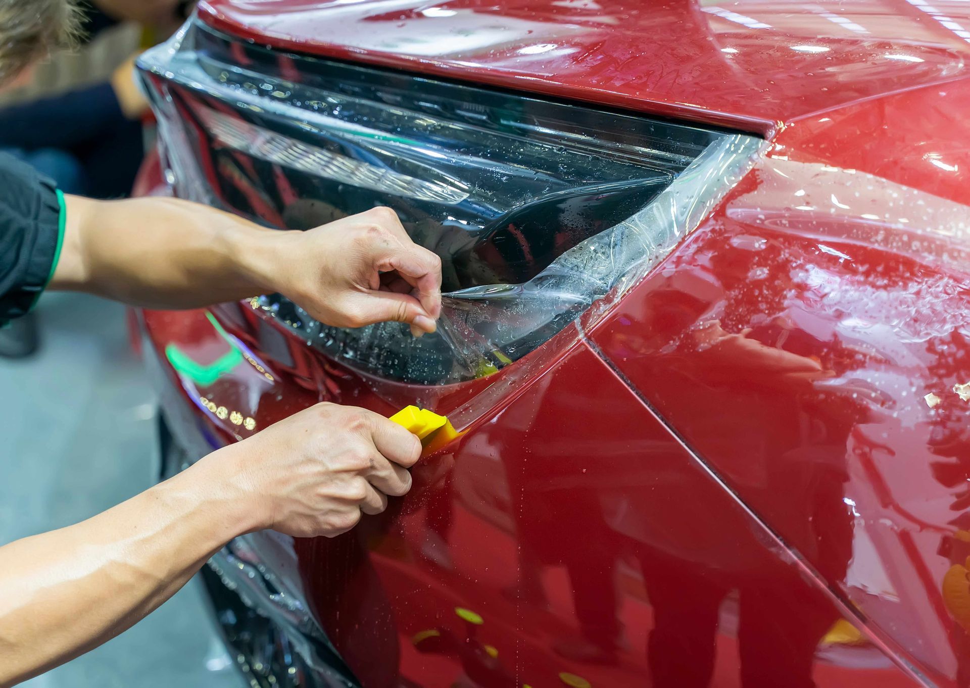 Hands applying protective film to a red car's taillight, using a squeegee in a brightly lit setting.